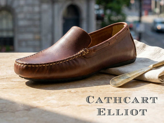 The Silverstone Men's Driving Loafer in Elk Pull-Up Leather by Orca Bay rests on a marble surface beside a beige cloth and shoehorn, with the Orca Bay logo at the bottom right and a blurred city street in the background.