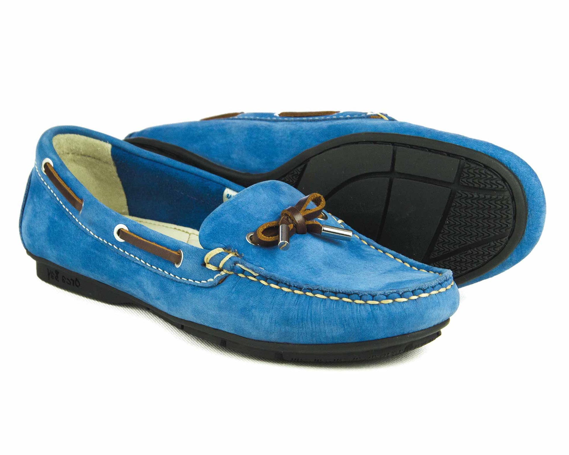 The BALLENA Ladies Powder Blue Suede Summer Loafer by Orca Bay combines style with comfort, featuring a decorative bow on top, brown leather lace accents, a rubber sole, and white stitching along the edges. These loafers make the perfect driving shoe for women.
