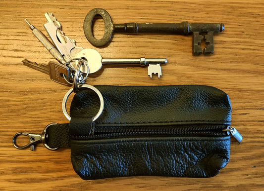 The Cathcart Elliot Black Grain Leather Key Wallet with zip and clip rests on a wooden surface, accompanied by several modern metal keys, a large vintage skeleton key, a tubular lock key, and a miniature key.