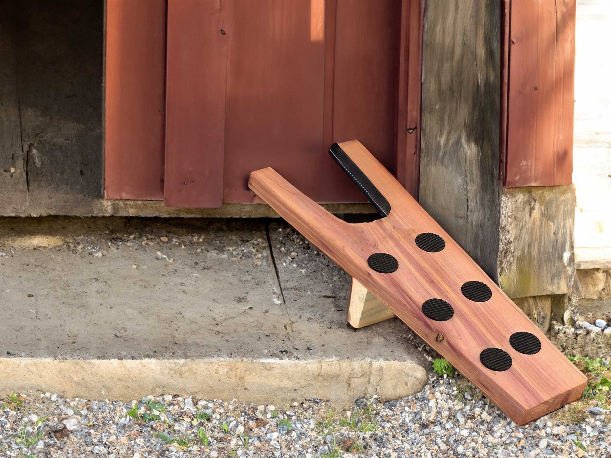 A wooden barn door ramp with six black non-slip circles rests against a barn entrance, partially open. The ramp leads from a gravel surface to the barns wooden floor.