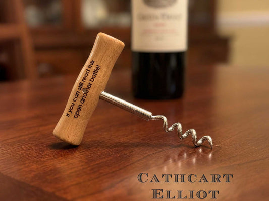 A Cathcart Elliot Personalised T Handle Corkscrew | Wooden Wine Opener, engraved with "If you can still read this, open another bottle," rests on a wooden table with a blurred wine bottle in the background.