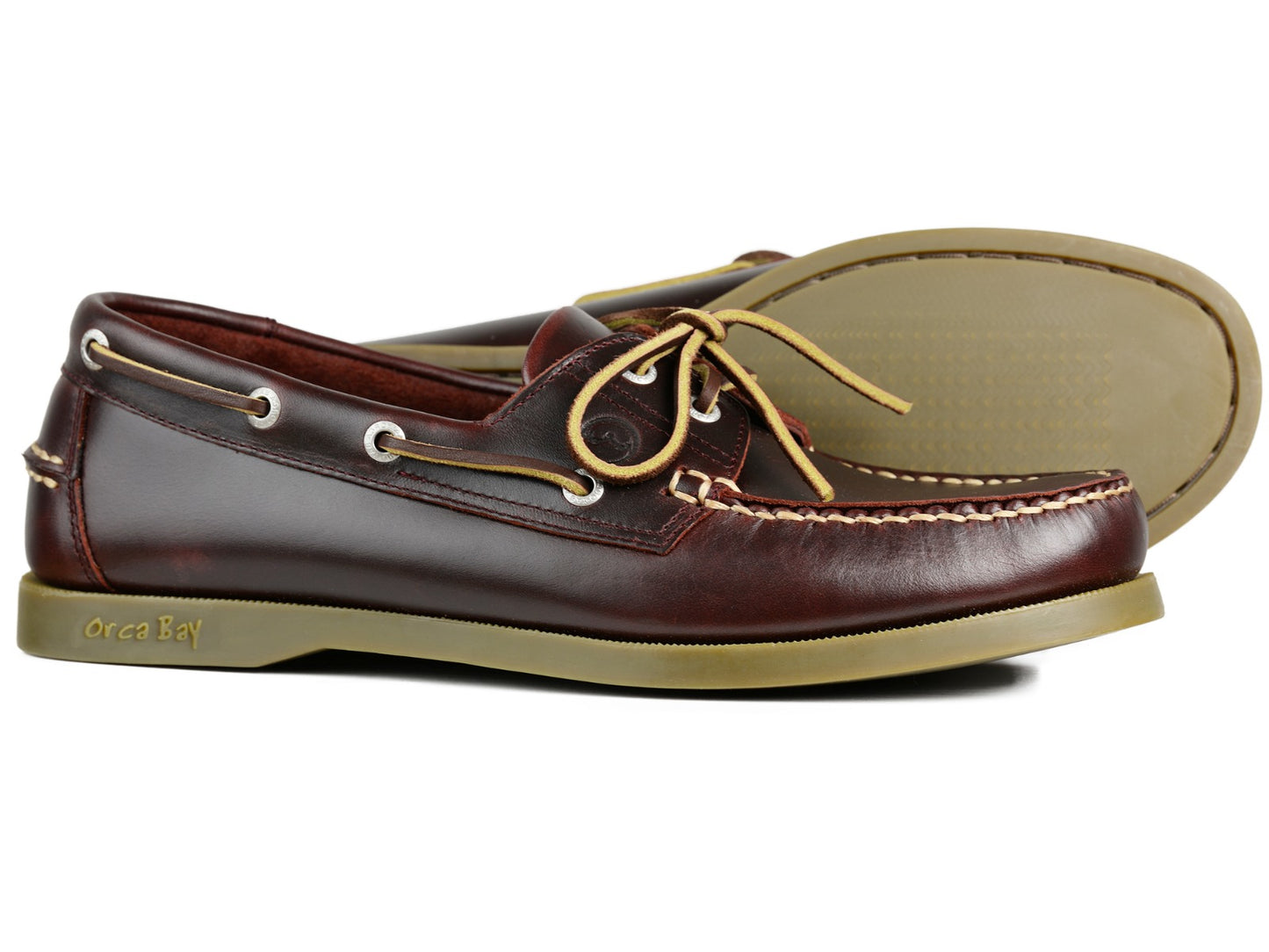 The CREEK Mens Burgundy leather deck shoes by Orca Bay feature premium stitching, slip-resistant soles, and beige laces. One shoe stands upright while the other lies on its side, allowing a view of the sole and the Orca Bay logo.