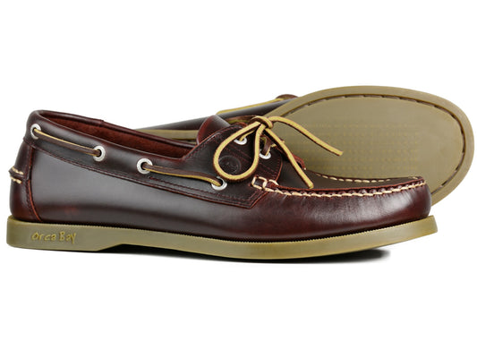 The CREEK Mens Burgundy leather deck shoes by Orca Bay feature premium stitching, slip-resistant soles, and beige laces. One shoe stands upright while the other lies on its side, allowing a view of the sole and the Orca Bay logo.