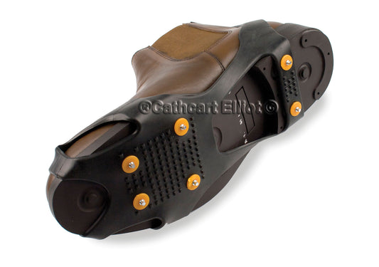 A close-up shows the sole of a brown shoe fitted with Cathcart Elliot’s Studded Ice Grippers, a black and orange winter traction aid with metal studs for enhanced grip on slippery surfaces.