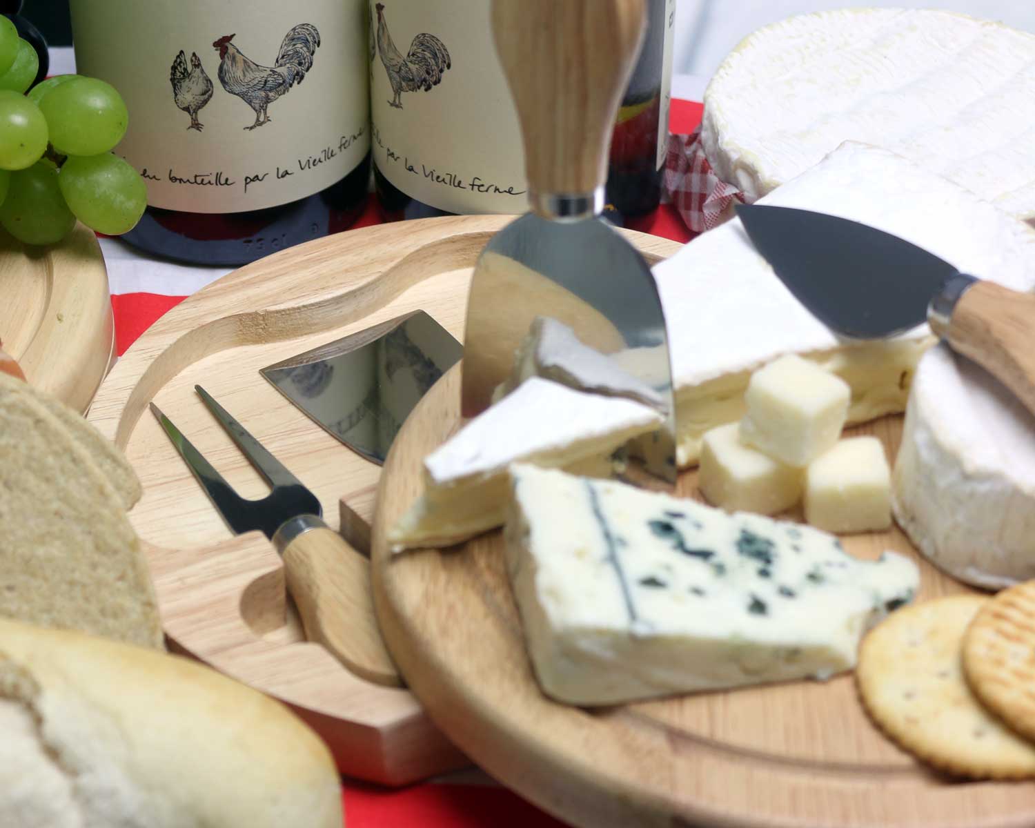 Cheese board with various cheeses, including blue cheese, Brie, and blocks of hard cheese. Surrounded by bread, grapes, crackers, and wooden-handled cheese knives. Bottles of wine with rooster labels in the background.