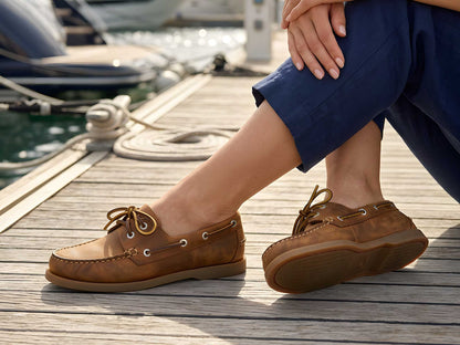 A person in navy pants sits on a wooden dock by the water, wearing Orca Bay Creek Sand Nubuck Women’s Boat Shoes from Cathcart Elliot. Their legs are crossed, with boats blurred in the background.