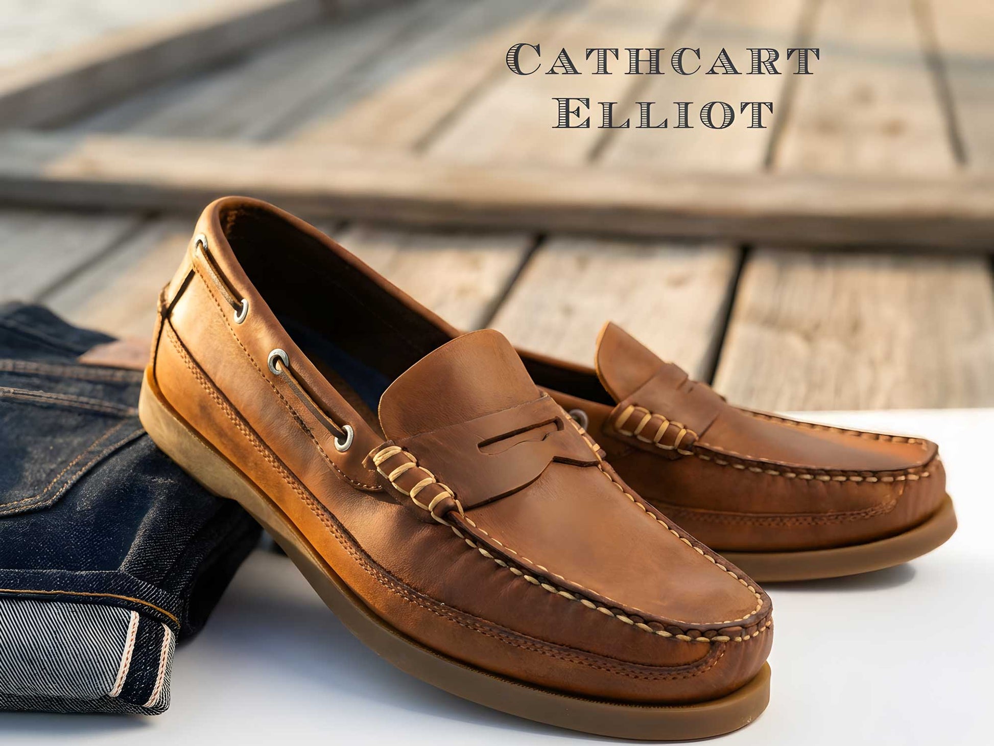 A pair of Orca Bay Fripp Sand Nubuck men's slip-on deck shoes sit on a wooden surface next to folded denim jeans, with "Cathcart Elliot" displayed in the background.
