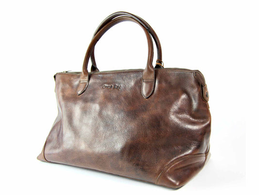The SALE MISSENDON Overnight by Orca Bay is a dark brown leather bag featuring stitched handles, zip closure, and an embossed logo. It offers a smooth texture with patterned fabric lining inside and a structured, elegant design outside.