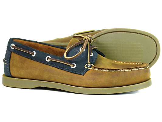 The OAKLAND Mens Sand Navy Nubuck Deck Shoes by Orca Bay feature nubuck in brown and navy with tan laces, white stitching, and beige razor-cut rubber soles for grip. One shoe stands upright while the other lies on its side, highlighting their stylish texture.
