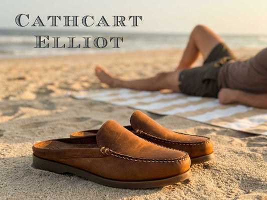 A pair of Orca Bay Mule Men’s Slip-On Deck Shoes in sand oiled nubuck rests on the sand, sharply in focus, while someone lounges on a striped beach towel by the ocean. The text “Cathcart Elliot” appears in the upper left corner.