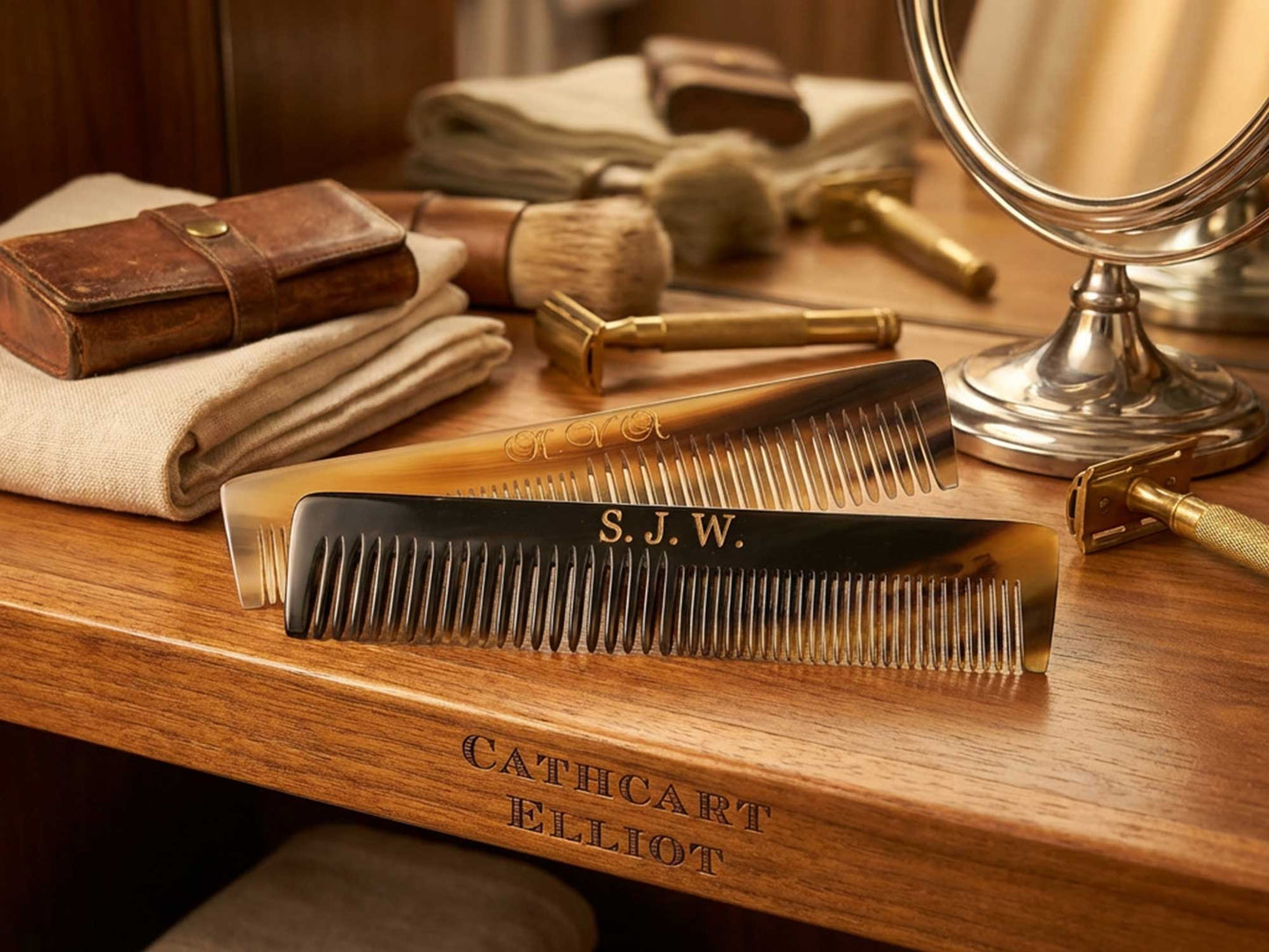 Two personalised 6-inch handmade real ox horn combs by Cathcart Elliot, one engraved with S.J.W., rest on a wooden vanity beside a mirror, towel, brushes, leather case, and have "Cathcart Elliot" elegantly engraved on the vanity.