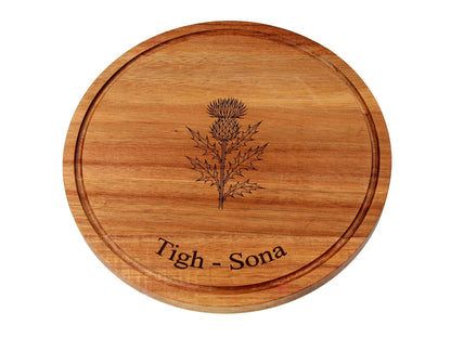 The Cathcart Elliot Personalised Round Acacia Wood Cheese and Cutting Board (25cm x 1.5cm) features a carved thistle and "Tigh - Sona" engraving—ideal as a unique serving board or sustainable cheese platter.