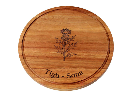 The Cathcart Elliot Personalised Round Acacia Wood Cheese and Cutting Board (25cm x 1.5cm) features a carved thistle and "Tigh - Sona" engraving—ideal as a unique serving board or sustainable cheese platter.