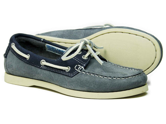 The SANDUSKY Mens Deck Shoe by Orca Bay, in indigo and grey, features rawhide laces and non-slip white soles. Visible stitching along the sides and toe area adds style. One shoe lies on its side, with the other propped up behind it.