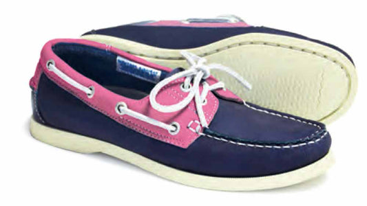A pair of Orca Bay SANDUSKY Ladies Deck Shoes in indigo blue and magenta pink, featuring white laces and non-slip soles. One shoe is on its side to display the sole, while the other stands upright, highlighting the classic design with stitching and side eyelet details.