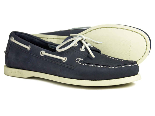 The SANDUSKY Mens Indigo Deck Shoe by Orca Bay features a pair of dark blue deck shoes with white laces and stitching. These casual shoes boast non-slip white rubber soles and include two eyelets on each side for lacing.