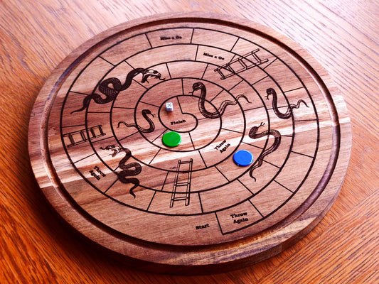 The Cathcart Elliot Personalised Acacia Wood Round Snakes & Ladders Game Board features engraved snakes, ladders, a die, and green and blue counters on wood—a unique gift with a spiral path leading to the Finish.