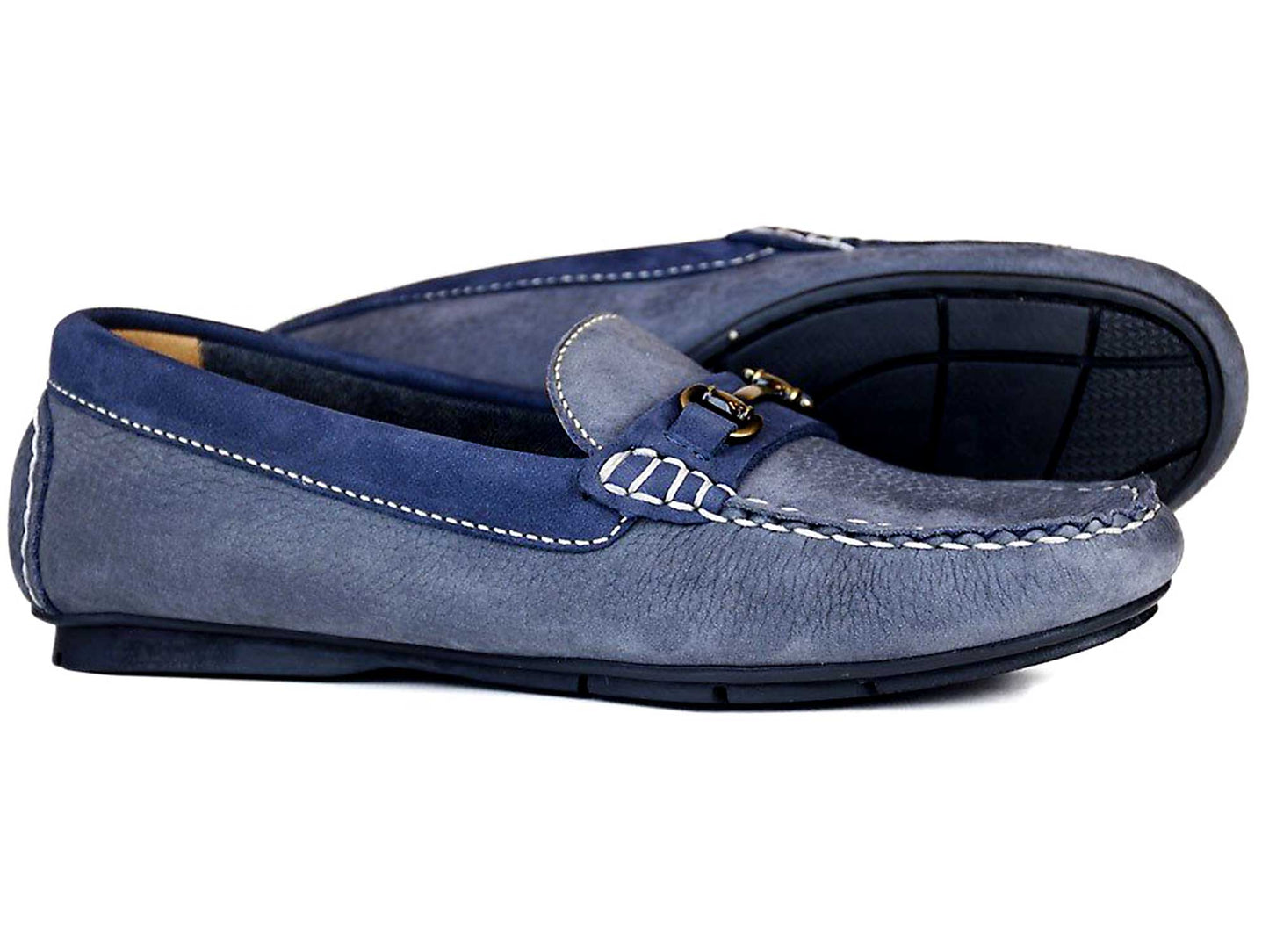 The Orca Bay VERONA Ladies Loafers in Grey Indigo Nubuck feature dark blue accents and a decorative metal bit on the front, with a stitched design and non-slip black rubber soles. One shoe is tilted to showcase its tread pattern underneath.