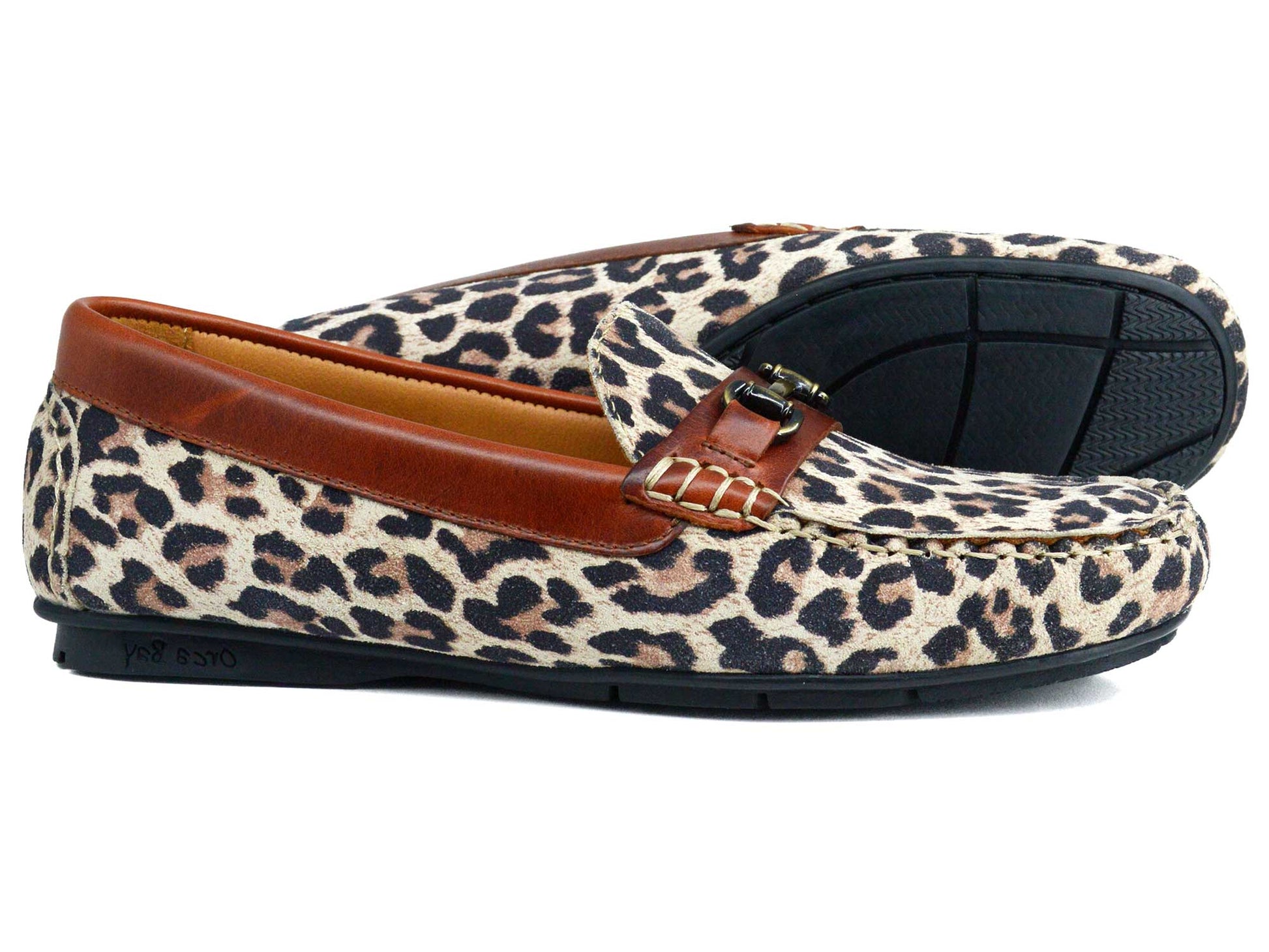 The Orca Bay VERONA Ladies Loafers feature a leopard print in premium suede, brown leather trim, a decorative buckle, and black non-slip soles. One shoe stands upright while the other lies on its side to show the sole.