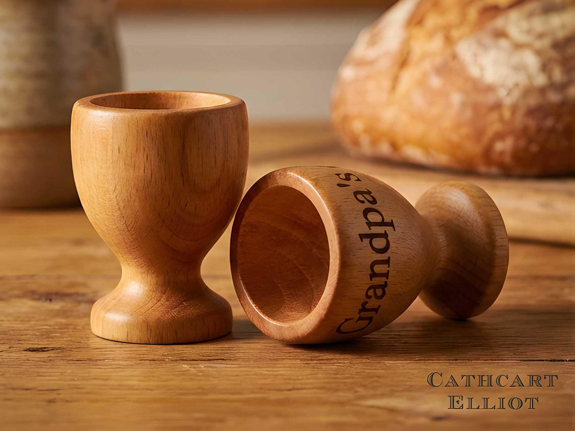Two beech egg cups by Cathcart Elliot rest on wood—one upright, one sideways with "Grandpa's" engraved. A blurred loaf of bread in the background draws focus to the personalised, laser-engraved set of 2.