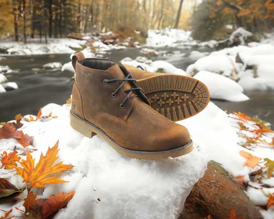 A pair of brown leather boots is displayed on a snowy rock beside a flowing river. Autumn leaves are scattered around, and trees with fall foliage are visible in the background.